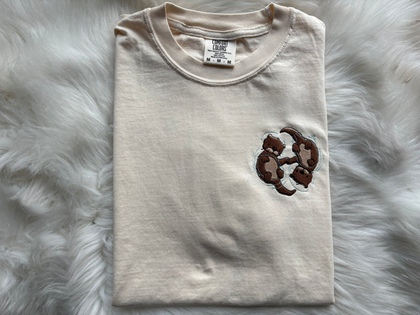 A cream coloured shirt on a fuzzy white background. The shirt has a small, pocket-sized image of two otters holding hands with blue water ripples around them.